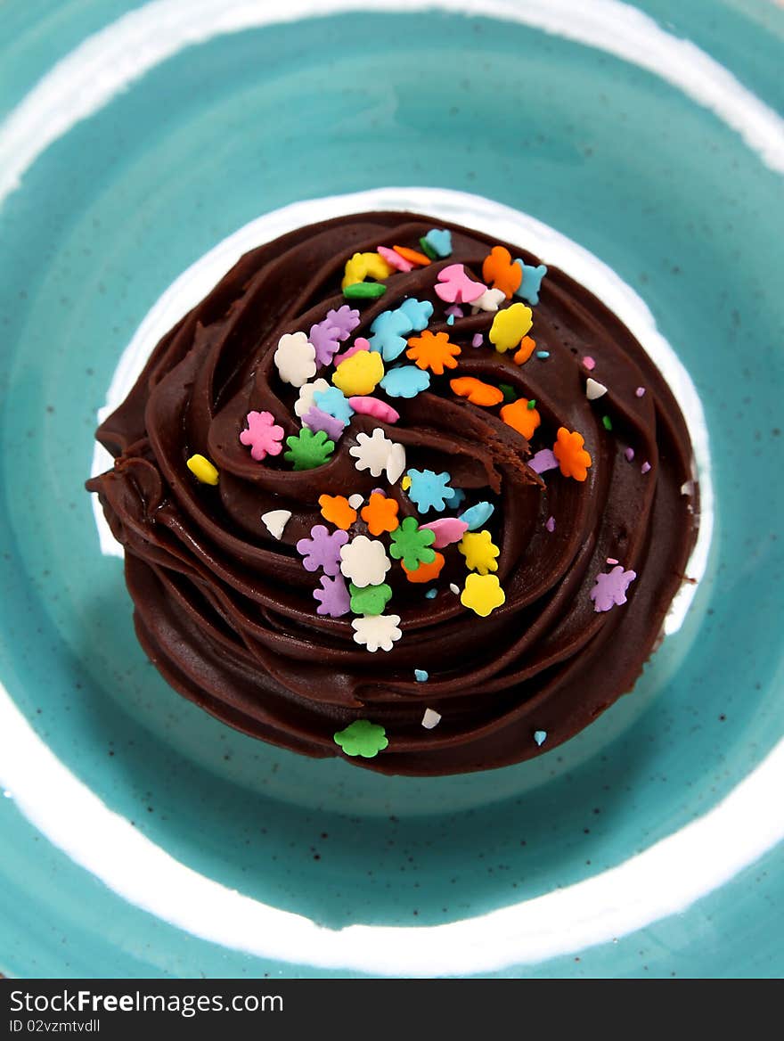 A delicious chocolate frosted cupcake with multicolored sprinkles sitting on a bright blue plate as seen from above. A delicious chocolate frosted cupcake with multicolored sprinkles sitting on a bright blue plate as seen from above