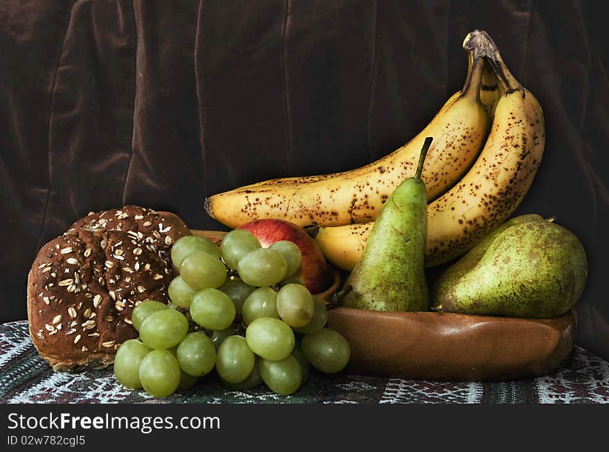 Classic still life with ripe fruit in a wooden bowl with breadroll - currant bun green grapes, ripe bunch of banana and green pears. Classic still life with ripe fruit in a wooden bowl with breadroll - currant bun green grapes, ripe bunch of banana and green pears