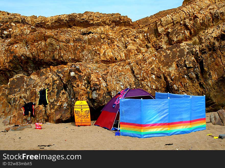 Colourful beach tent and windbreaker with wetsuits, bucket and spade and a body board on a deserted Devon beach in the UK with a lovely rock formation background. Colourful beach tent and windbreaker with wetsuits, bucket and spade and a body board on a deserted Devon beach in the UK with a lovely rock formation background.