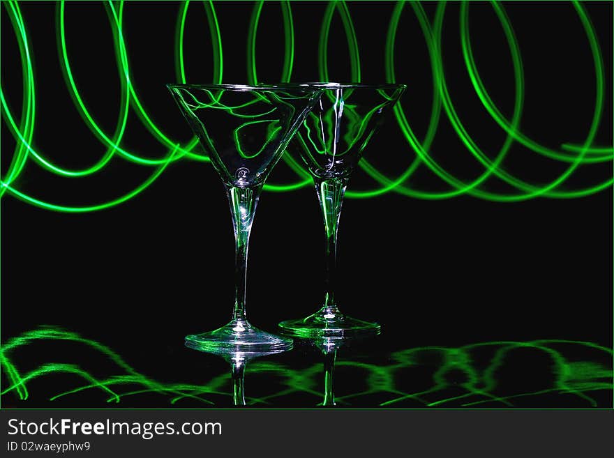 Pair martini glasses with black background and reflective black surface, lit with green led light trails. Pair martini glasses with black background and reflective black surface, lit with green led light trails