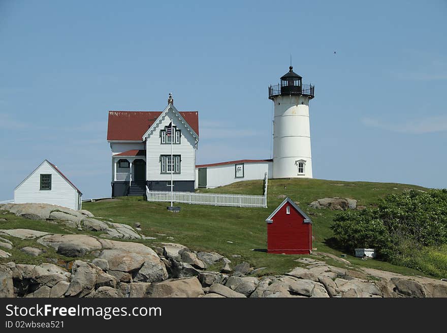 A lighthouse with out-buildings perched on a rocky landscape with green yard and blue, clear sky. A lighthouse with out-buildings perched on a rocky landscape with green yard and blue, clear sky.