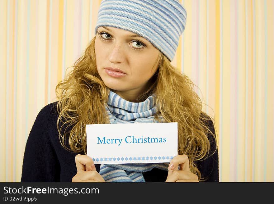 Young beautiful woman in a sweater with a blue hat and scarf holding a card with the text Merry Christmas. Young beautiful woman in a sweater with a blue hat and scarf holding a card with the text Merry Christmas