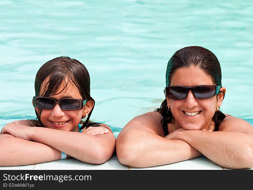 Daughter and mother smiling and wearing sunglasses at the border of a swimming pool. Daughter and mother smiling and wearing sunglasses at the border of a swimming pool