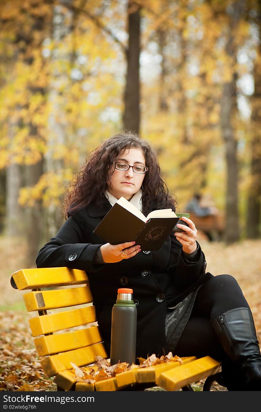 Young woman reading book in autumn park on bench