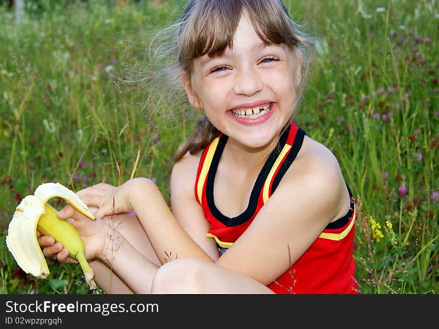 The joyful girl sits on a green glade eats a banana and laughs. The joyful girl sits on a green glade eats a banana and laughs