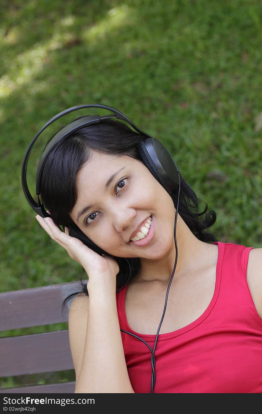 A smiling Asian woman listening to and enjoying the music on the headphone while relaxing in the park. A smiling Asian woman listening to and enjoying the music on the headphone while relaxing in the park