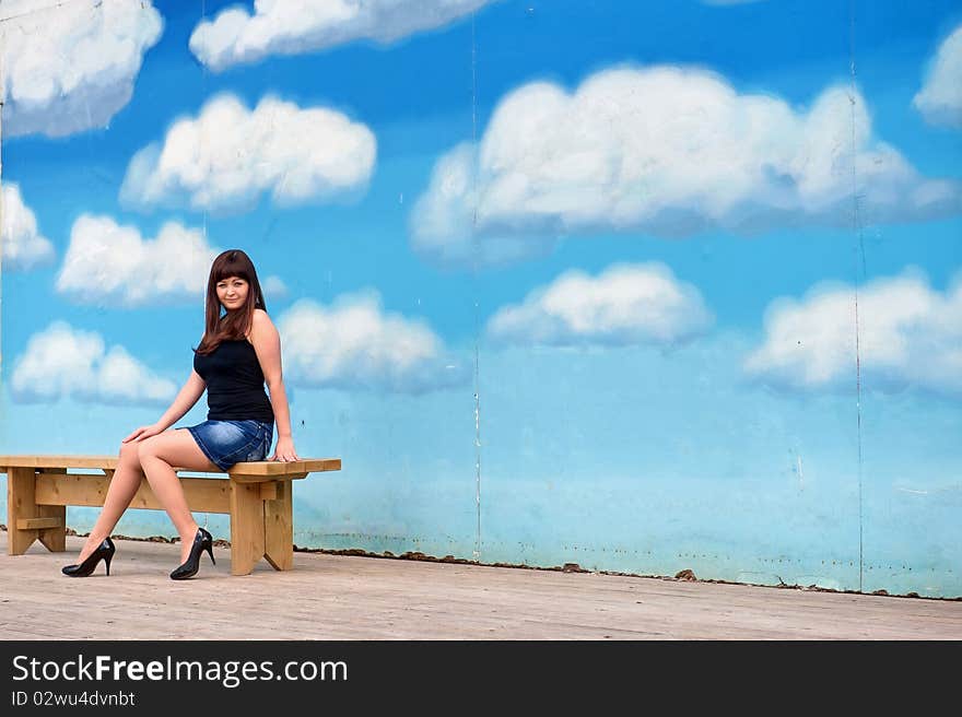 Young, woman in short skirt sit on the bench on the background of blue sky drawing. Young, woman in short skirt sit on the bench on the background of blue sky drawing.