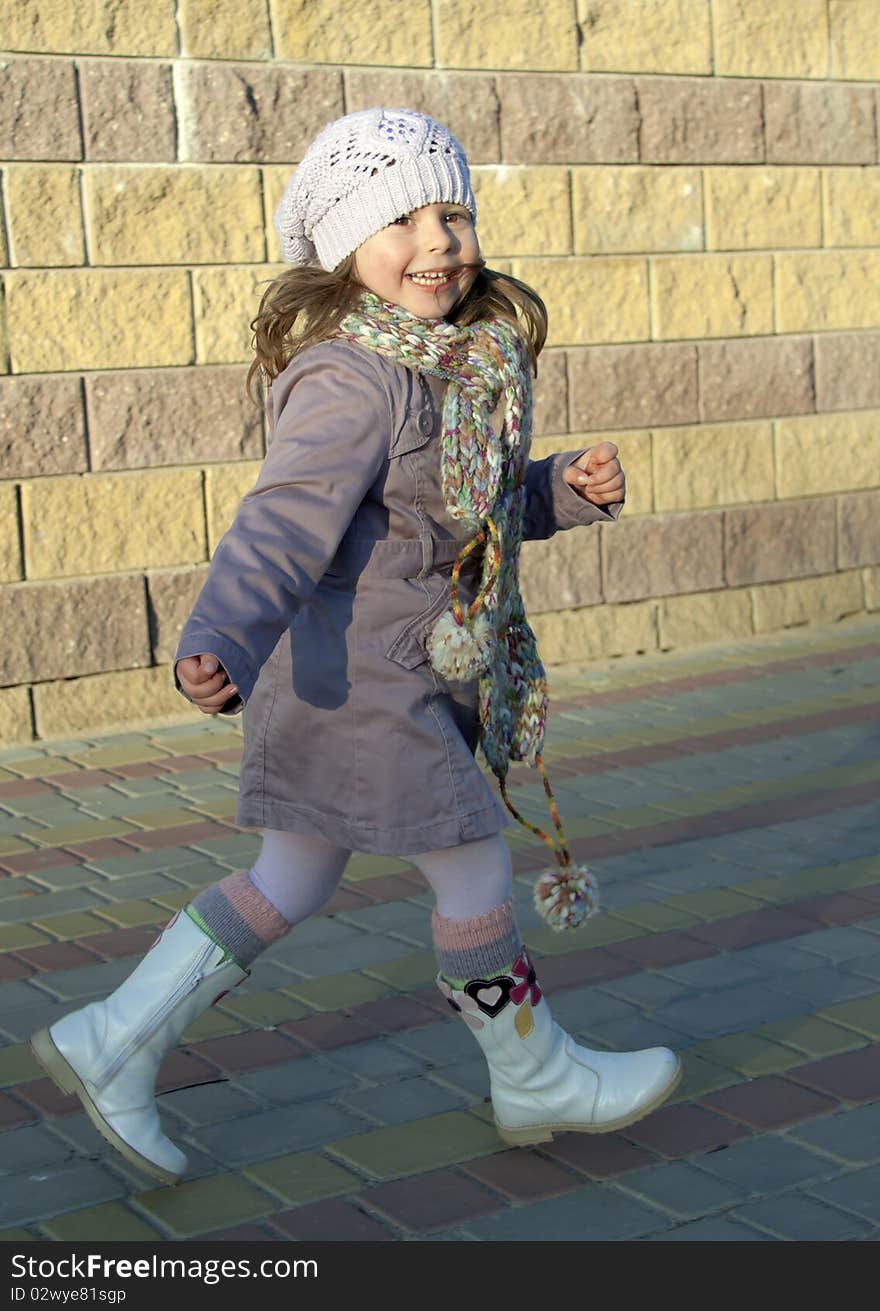 The joyful child in a raincoat with a scarf and boots runs along a stone wall. The joyful child in a raincoat with a scarf and boots runs along a stone wall