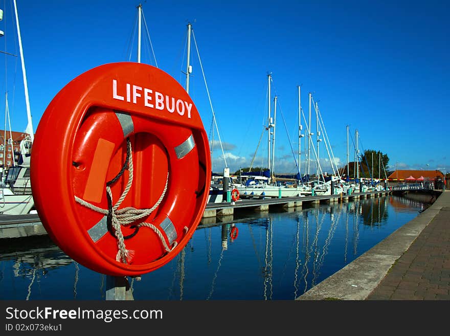 Bright orange lifebuoy captured on a bright sunny day with a deep blue sky on Weymouth Marina, with yachts in the background and reflections in the almost still waters. Bright orange lifebuoy captured on a bright sunny day with a deep blue sky on Weymouth Marina, with yachts in the background and reflections in the almost still waters.