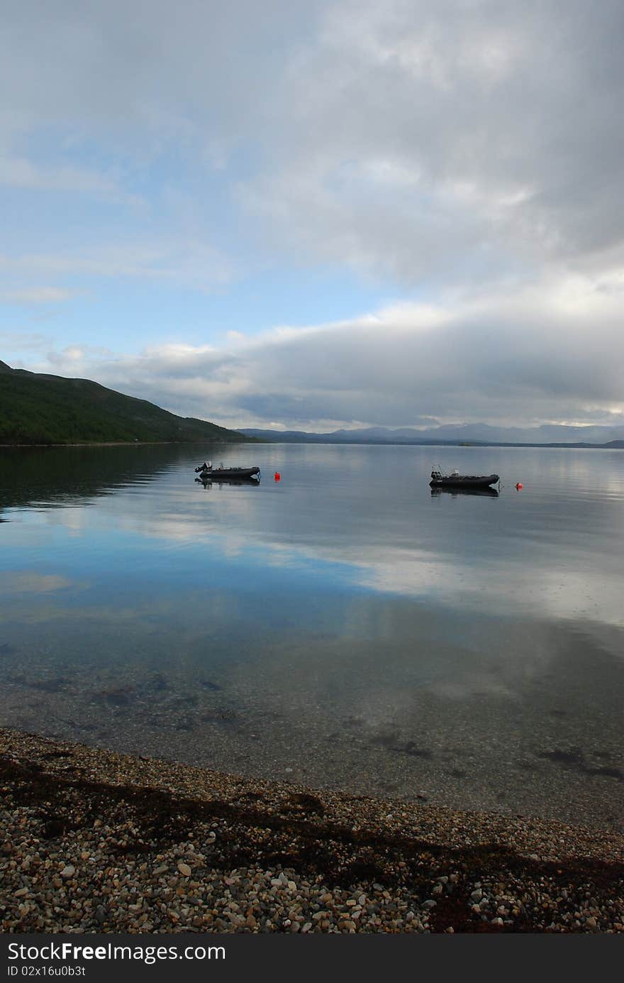 Stones and transparante water are at foreground and sun light crossinf frog and clouds and refrecting in ware surfce, two motorboats are in mirrow surface of water. Stones and transparante water are at foreground and sun light crossinf frog and clouds and refrecting in ware surfce, two motorboats are in mirrow surface of water