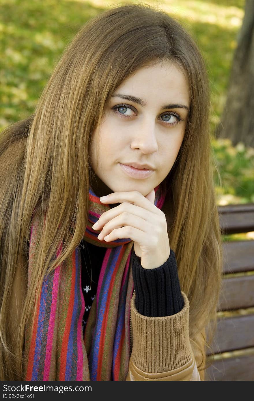 Vertical portrait of a beautiful young woman sitting on a bench in the park around a lot of yellow, red green leaves. Rests his head on his hand. Vertical portrait of a beautiful young woman sitting on a bench in the park around a lot of yellow, red green leaves. Rests his head on his hand