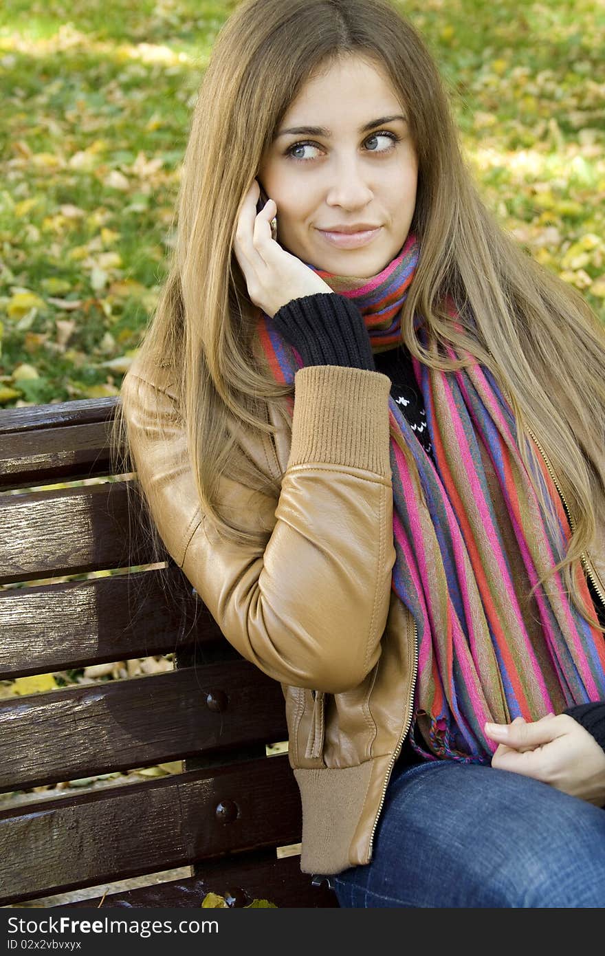 Young Caucasian woman calls on a mobile phone, sitting in a park on a wooden bench. Autumn around a lot of colorful foliage. Young Caucasian woman calls on a mobile phone, sitting in a park on a wooden bench. Autumn around a lot of colorful foliage