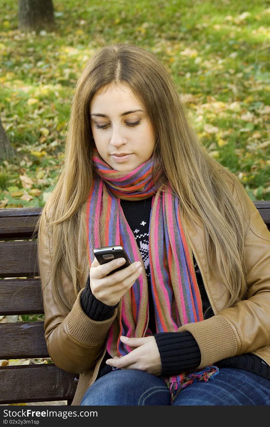 Young Caucasian woman with a cell phone, sitting in a park on a wooden bench, reading a SMS. Autumn around a lot of colorful foliage. Young Caucasian woman with a cell phone, sitting in a park on a wooden bench, reading a SMS. Autumn around a lot of colorful foliage