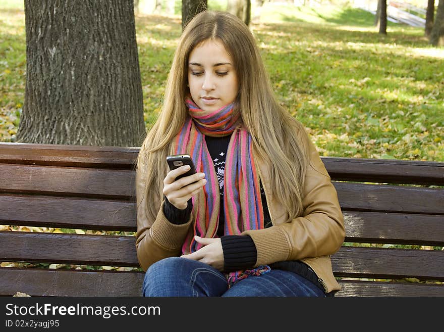 Young Caucasian woman with a cell phone, sitting in a park on a wooden bench, reading a SMS. Autumn around a lot of colorful foliage. Young Caucasian woman with a cell phone, sitting in a park on a wooden bench, reading a SMS. Autumn around a lot of colorful foliage