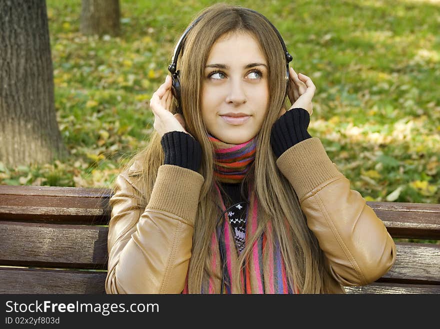 Young Caucasian woman with headphones in autumn park sitting on a wooden bench. Autumn around a lot of colorful foliage. Young Caucasian woman with headphones in autumn park sitting on a wooden bench. Autumn around a lot of colorful foliage