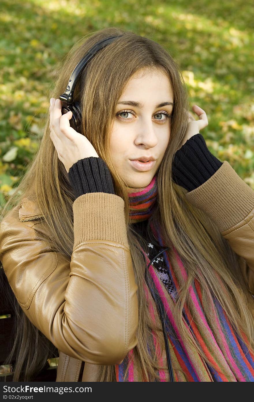 Young Caucasian woman with headphones in autumn park sitting on a wooden bench, listening to music and singing. Autumn around a lot of colorful foliage. Young Caucasian woman with headphones in autumn park sitting on a wooden bench, listening to music and singing. Autumn around a lot of colorful foliage