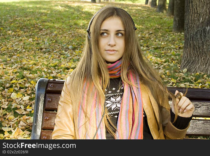 Young Caucasian woman with headphones in autumn park sitting on a wooden bench. Autumn around a lot of colorful foliage. Young Caucasian woman with headphones in autumn park sitting on a wooden bench. Autumn around a lot of colorful foliage