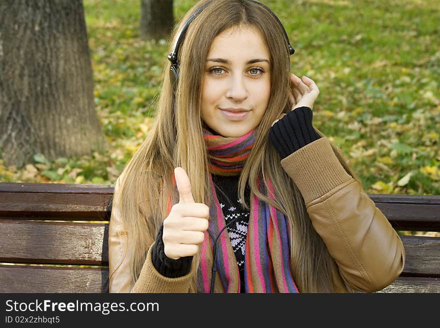 Young Caucasian woman with headphones in autumn park sitting on a wooden bench, listening to music. Thumbs up. Autumn around a lot of colorful foliage. Young Caucasian woman with headphones in autumn park sitting on a wooden bench, listening to music. Thumbs up. Autumn around a lot of colorful foliage