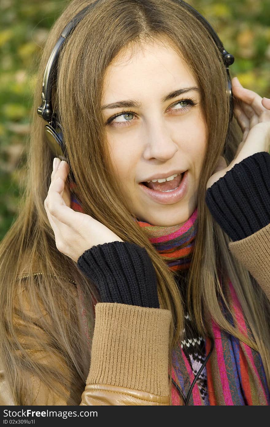 Young Caucasian woman with headphones in autumn park sitting on a wooden bench, listening to music and singing. Autumn around a lot of colorful foliage. Young Caucasian woman with headphones in autumn park sitting on a wooden bench, listening to music and singing. Autumn around a lot of colorful foliage