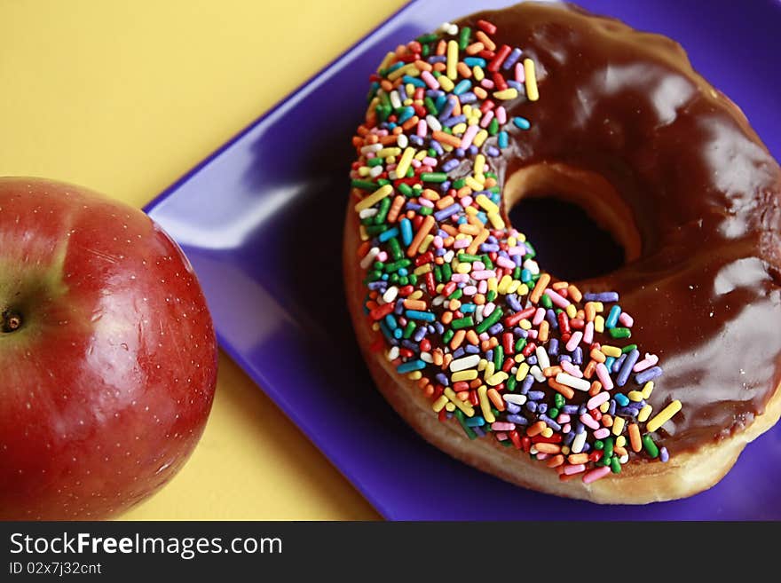 Chocolate glazed doughnut with rainbow sprinkles on plate with apple. Chocolate glazed doughnut with rainbow sprinkles on plate with apple