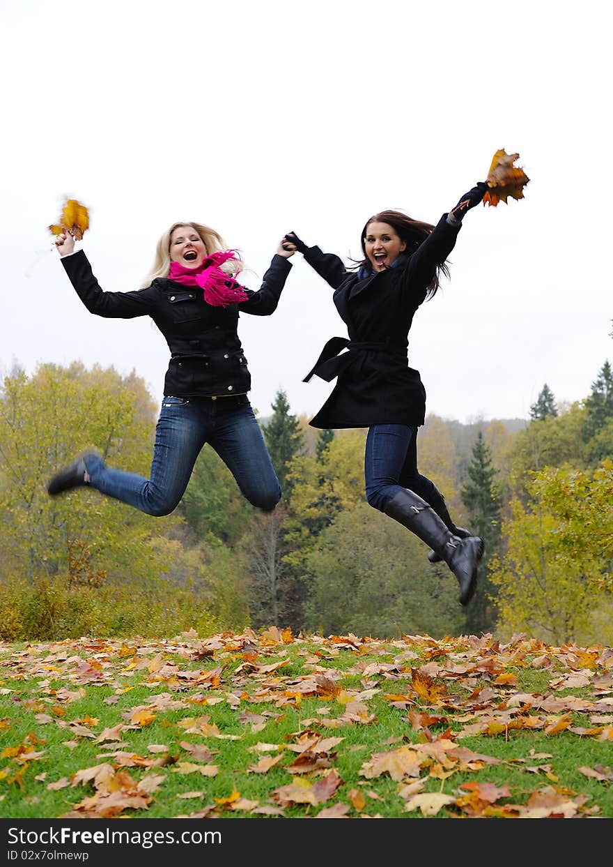 Two beautiful girl friends with autumn leafs in a park jumping. Two beautiful girl friends with autumn leafs in a park jumping