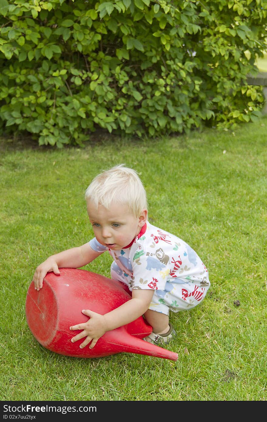 Cute blond toddler trying to pick up a red watering can. Cute blond toddler trying to pick up a red watering can