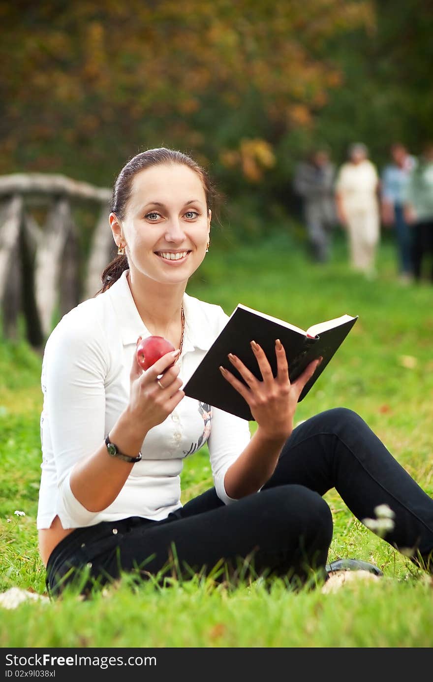 Youn smiling woman sitting on green grass with book and apple