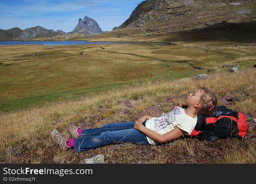 Small girl in focus, repose after walk, had on backpack, at Anayet plateau in Aragon, Spain. the lake and Beautiful peak du Midi d'Ossau in French site of Pytenees is at background. Small girl in focus, repose after walk, had on backpack, at Anayet plateau in Aragon, Spain. the lake and Beautiful peak du Midi d'Ossau in French site of Pytenees is at background