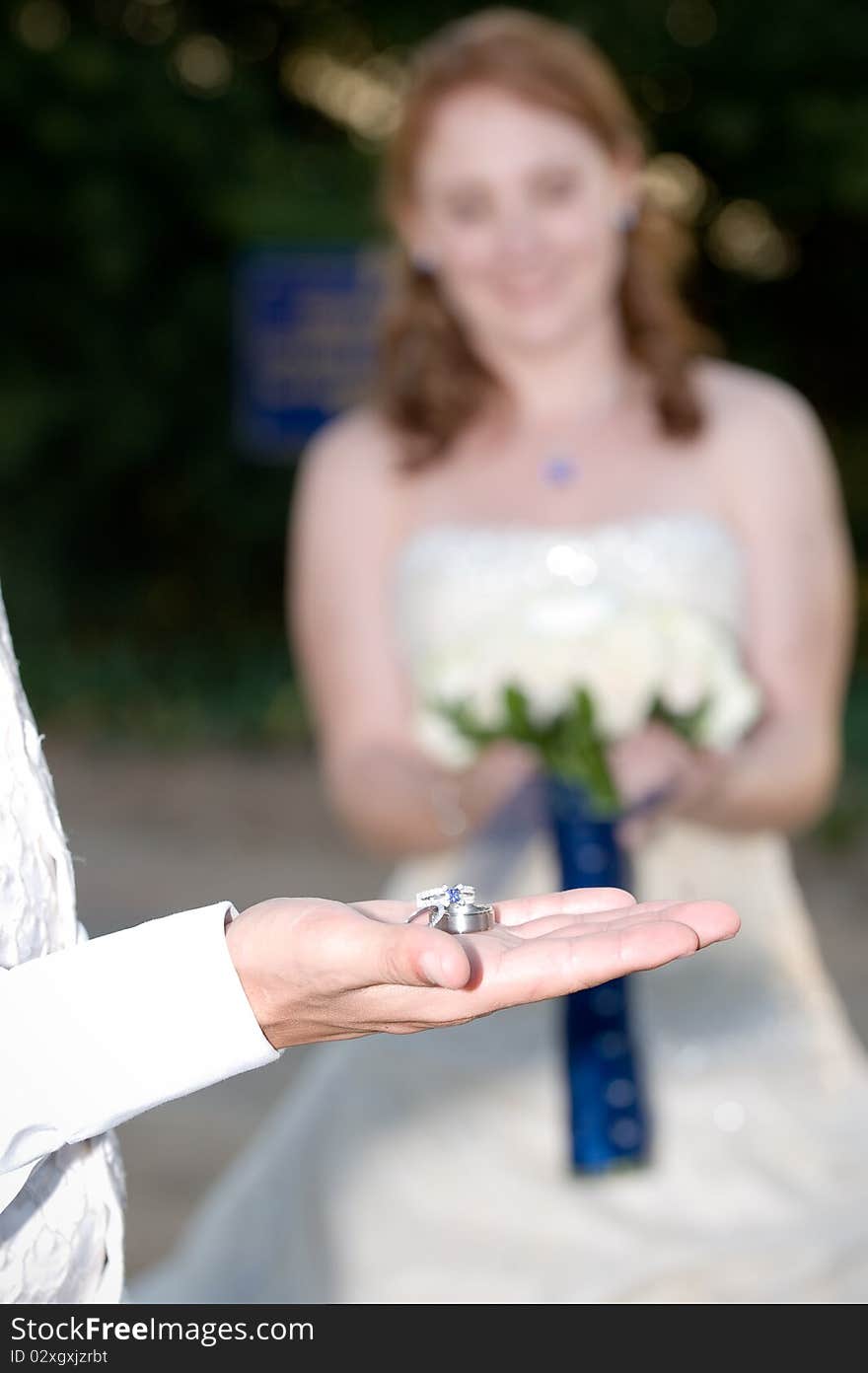 Wedding rings on the grooms palm with the bride smiling and holding flowers in the background