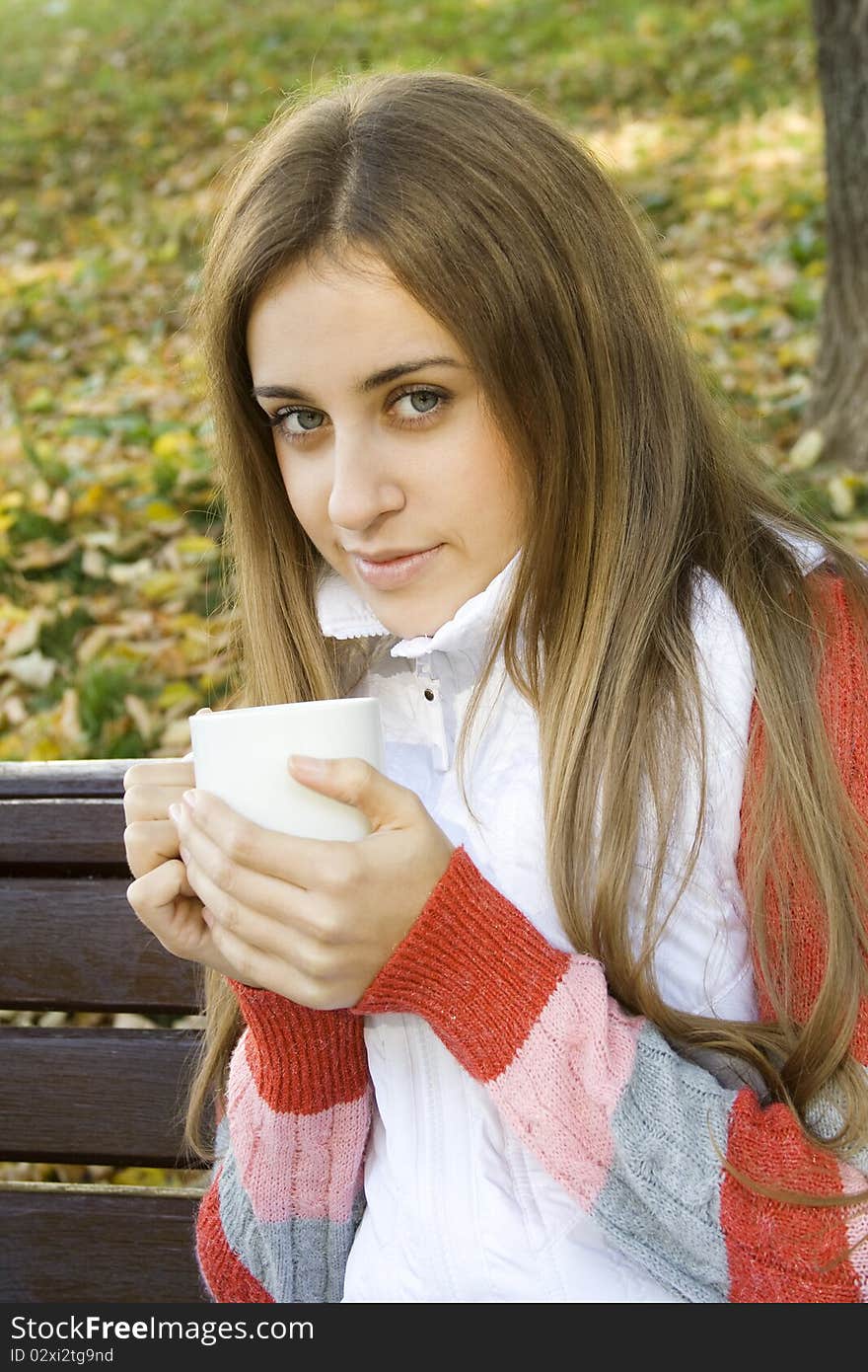 Beautiful young woman in autumn park sitting on a bench in the hands of white cup with coffee / tea. Beautiful young woman in autumn park sitting on a bench in the hands of white cup with coffee / tea