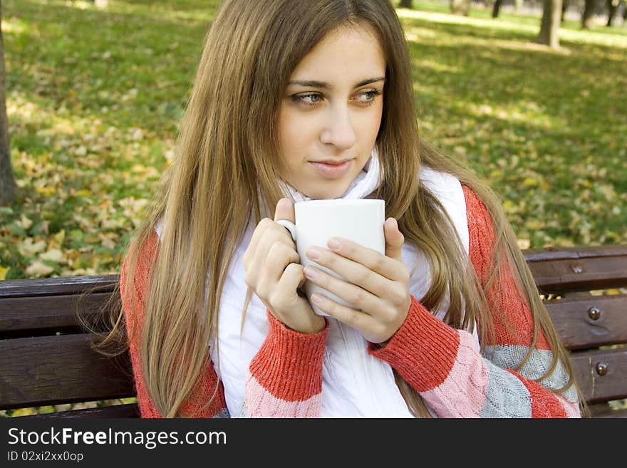 Beautiful young woman in autumn park sitting on a bench in the hands of white cup with coffee / tea. Beautiful young woman in autumn park sitting on a bench in the hands of white cup with coffee / tea