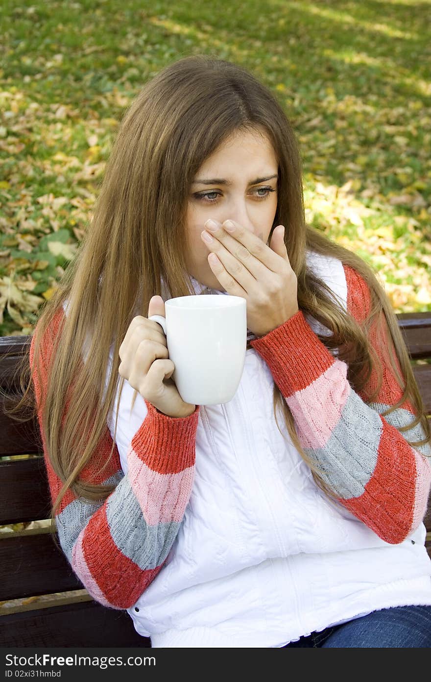 Beautiful young woman in autumn park sitting on a bench in the hands of white cup with coffee / tea. Beautiful young woman in autumn park sitting on a bench in the hands of white cup with coffee / tea