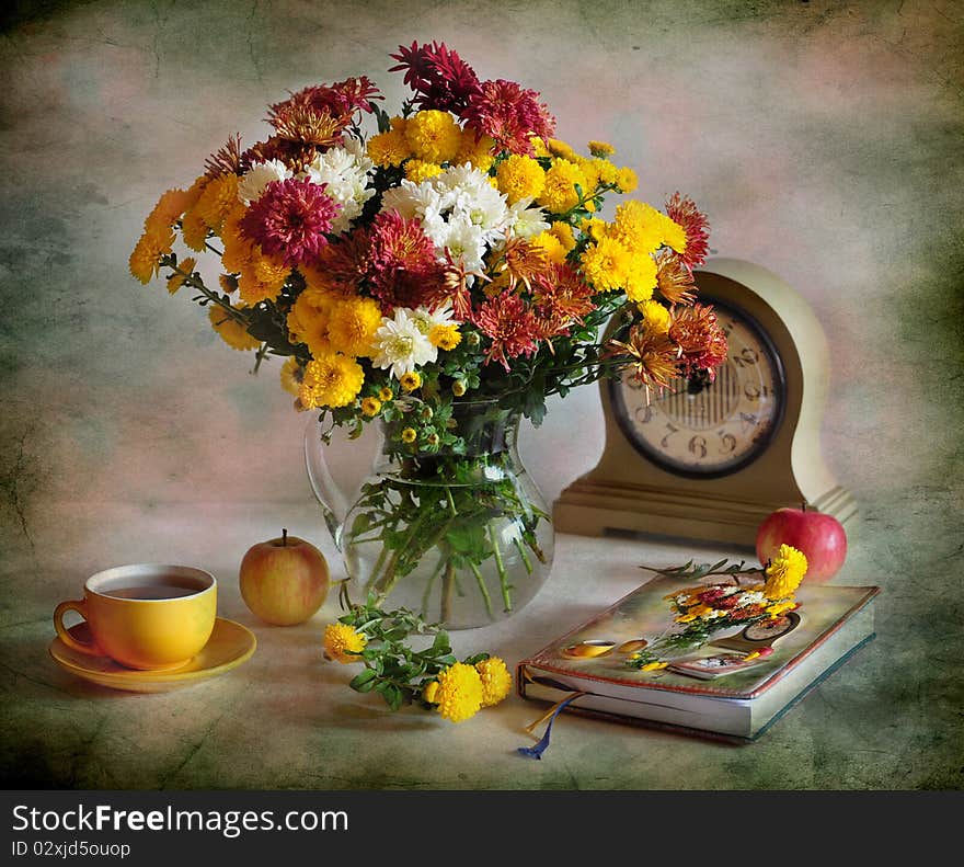 A bouquet of varicoloured chrysanthemums is in a glass jug, alongside apples, cup of tea, clock and notebook. A bouquet of varicoloured chrysanthemums is in a glass jug, alongside apples, cup of tea, clock and notebook