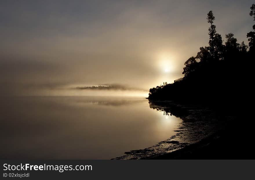 The sun rises over a misty Lake Paringo, on the West Coast of New Zealand. Drees can just be seen through the morning fog. The sun rises over a misty Lake Paringo, on the West Coast of New Zealand. Drees can just be seen through the morning fog.