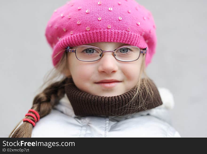 Adorable small girl in bright pink hat with long dark hair and glasses on grey background