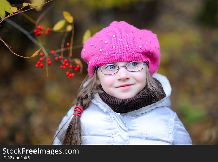 Adorable small girl in bright pink hat with long dark hair and glasses on beauty autumn background with rowanberry on it