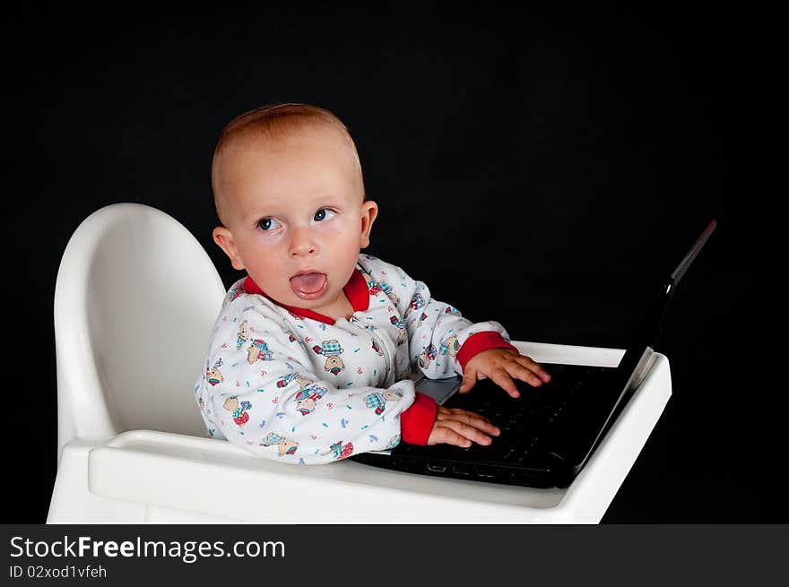 Baby sitting in the high chair, sticking out his tongue and using the laptop. Baby sitting in the high chair, sticking out his tongue and using the laptop