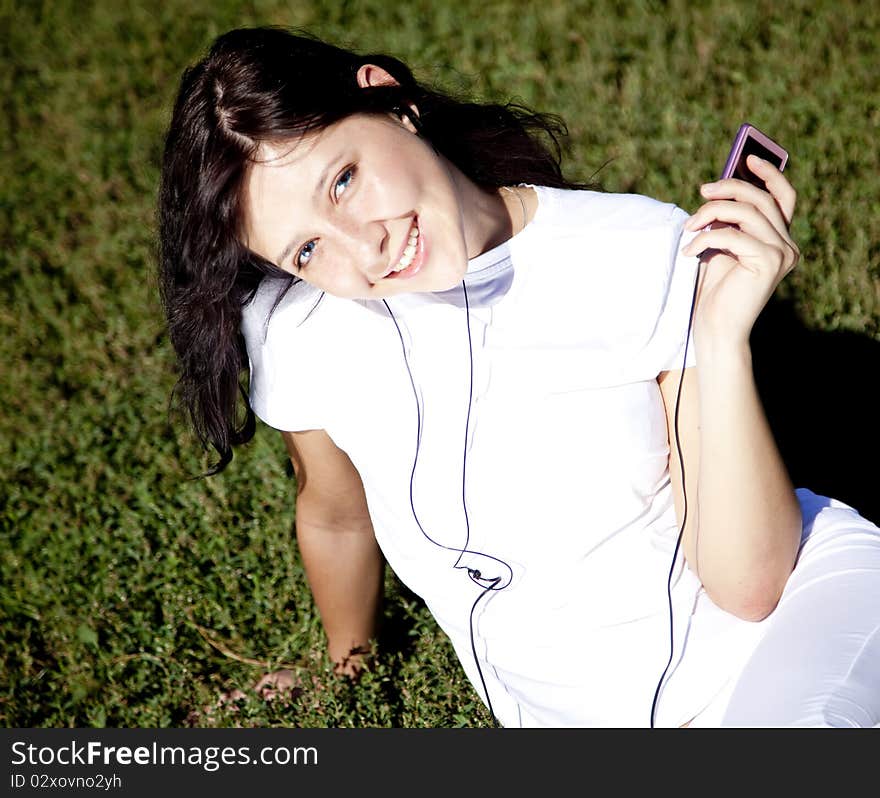 Pretty young brunette girl listening music on green grass in the park and show pink music player. Pretty young brunette girl listening music on green grass in the park and show pink music player.