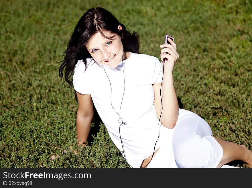 Pretty young brunette girl listening music on green grass in the park and show pink music player. Pretty young brunette girl listening music on green grass in the park and show pink music player.