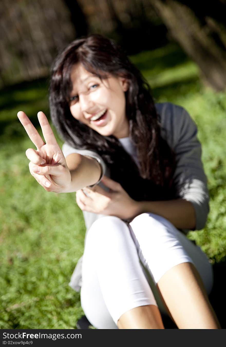 Portrait of beautiful brunette girl with blue eyes on green grass in the park show V symbol. Portrait of beautiful brunette girl with blue eyes on green grass in the park show V symbol.