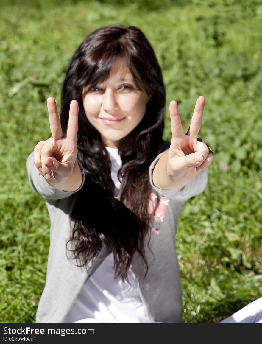 Portrait of beautiful brunette girl with blue eyes on green grass in the park show V symbol. Portrait of beautiful brunette girl with blue eyes on green grass in the park show V symbol.