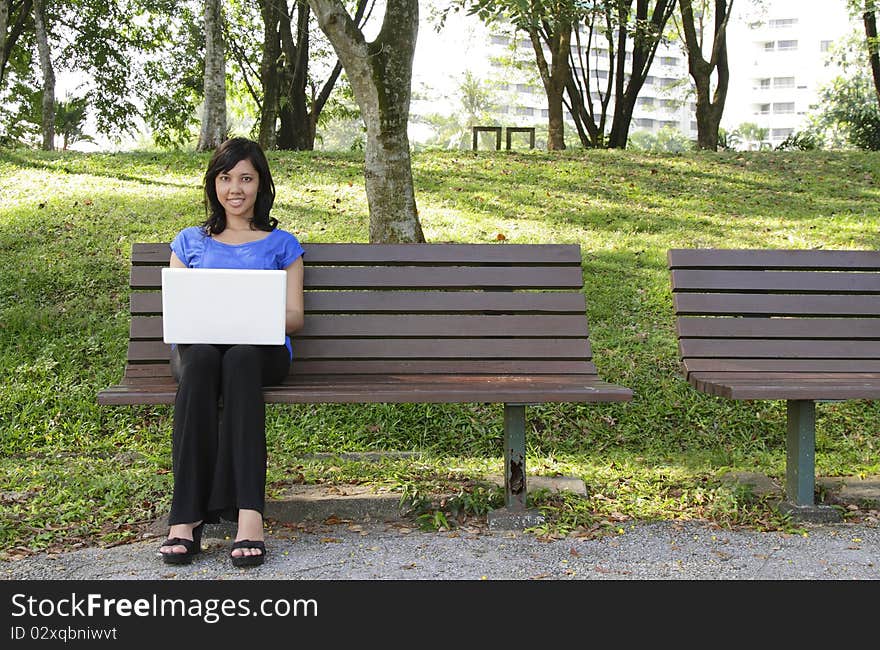 An Asian woman with a laptop sitting on a bench at a park. An Asian woman with a laptop sitting on a bench at a park