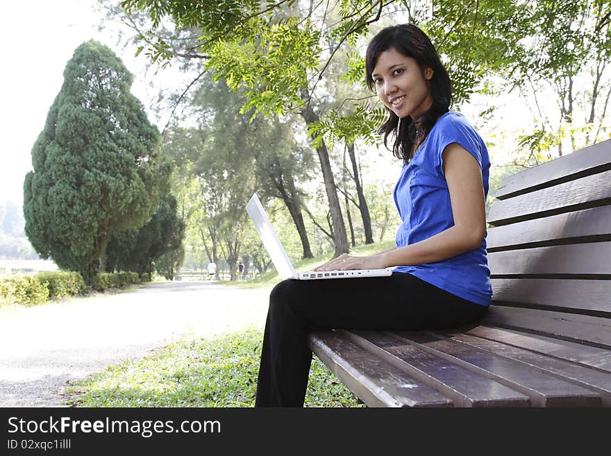 An Asian woman with a laptop sitting on a bench at a park. An Asian woman with a laptop sitting on a bench at a park