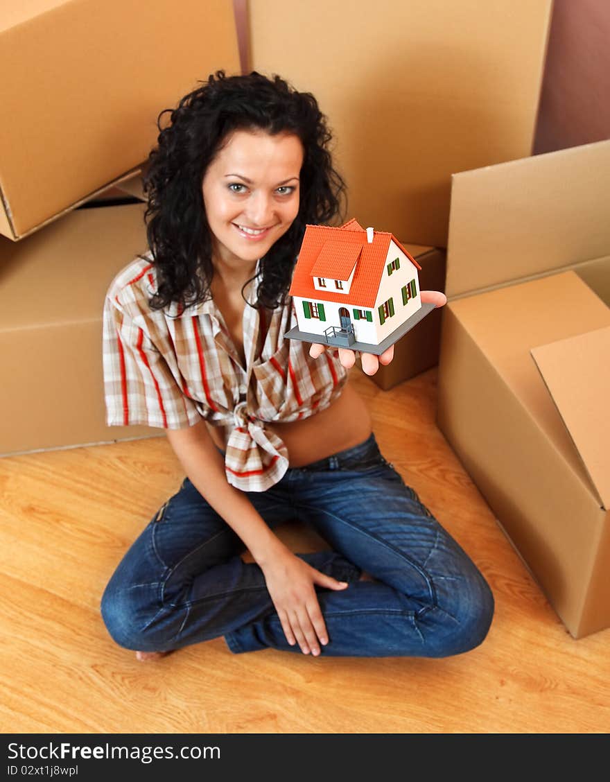 Attractive young woman offering a miniature house, boxes in the background. Attractive young woman offering a miniature house, boxes in the background