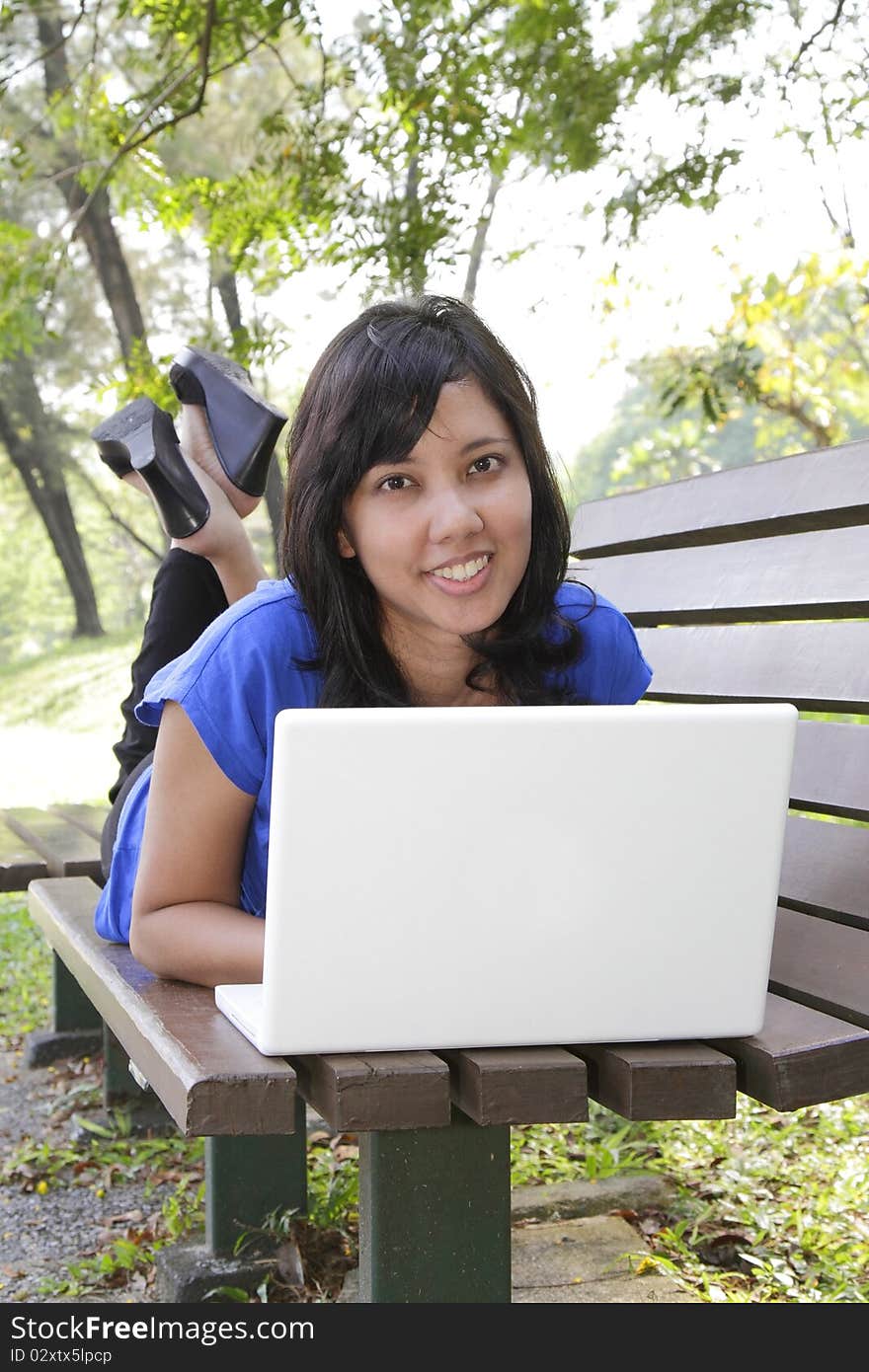 An Asian woman lying down on a bench using her laptop. An Asian woman lying down on a bench using her laptop