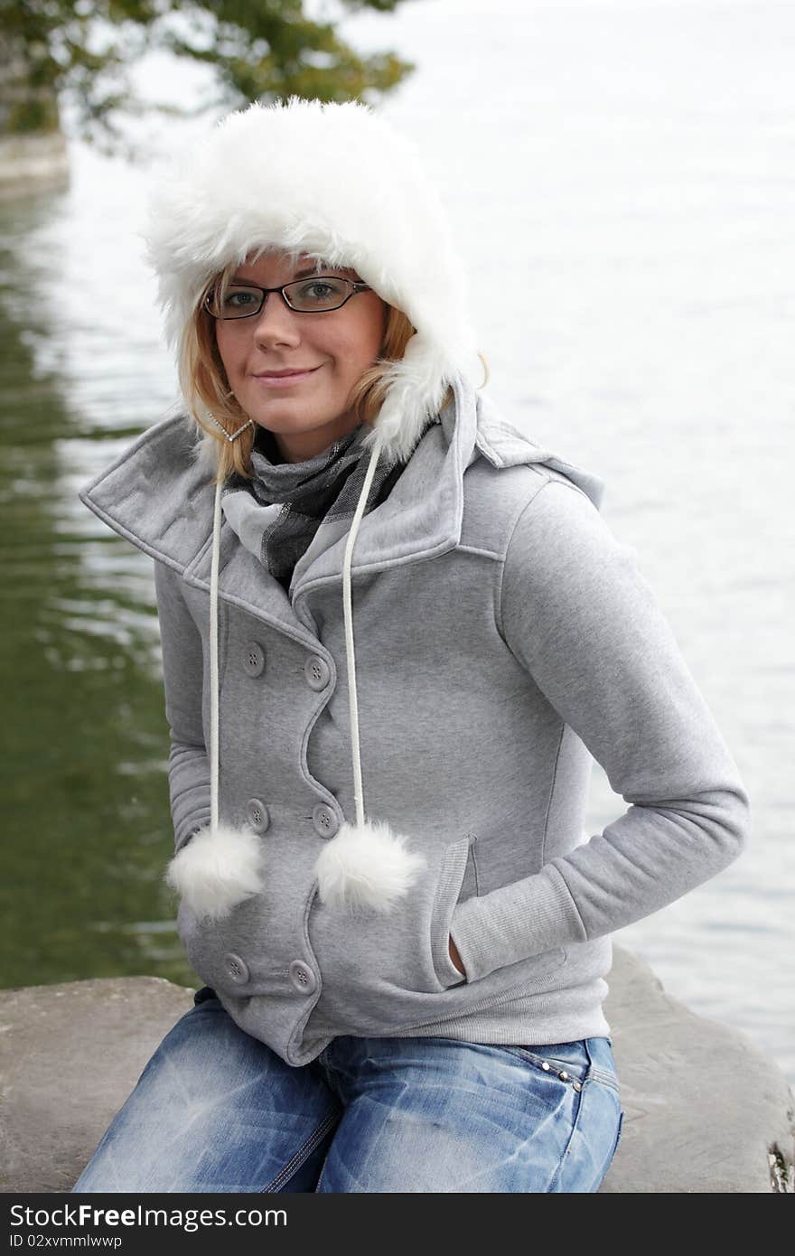 Young woman with white fur hat, smiling friendly looking into the camera. Autumn, monochrome mood at the lakeside. Young woman with white fur hat, smiling friendly looking into the camera. Autumn, monochrome mood at the lakeside.