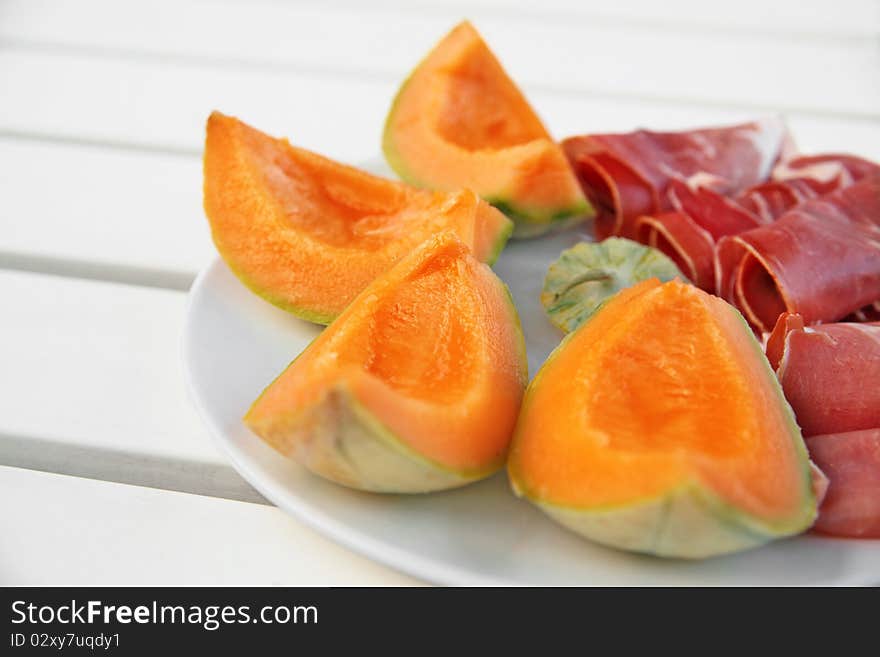 Slices of melon with italian ham in plain white plate on white picnic table. Slices of melon with italian ham in plain white plate on white picnic table.