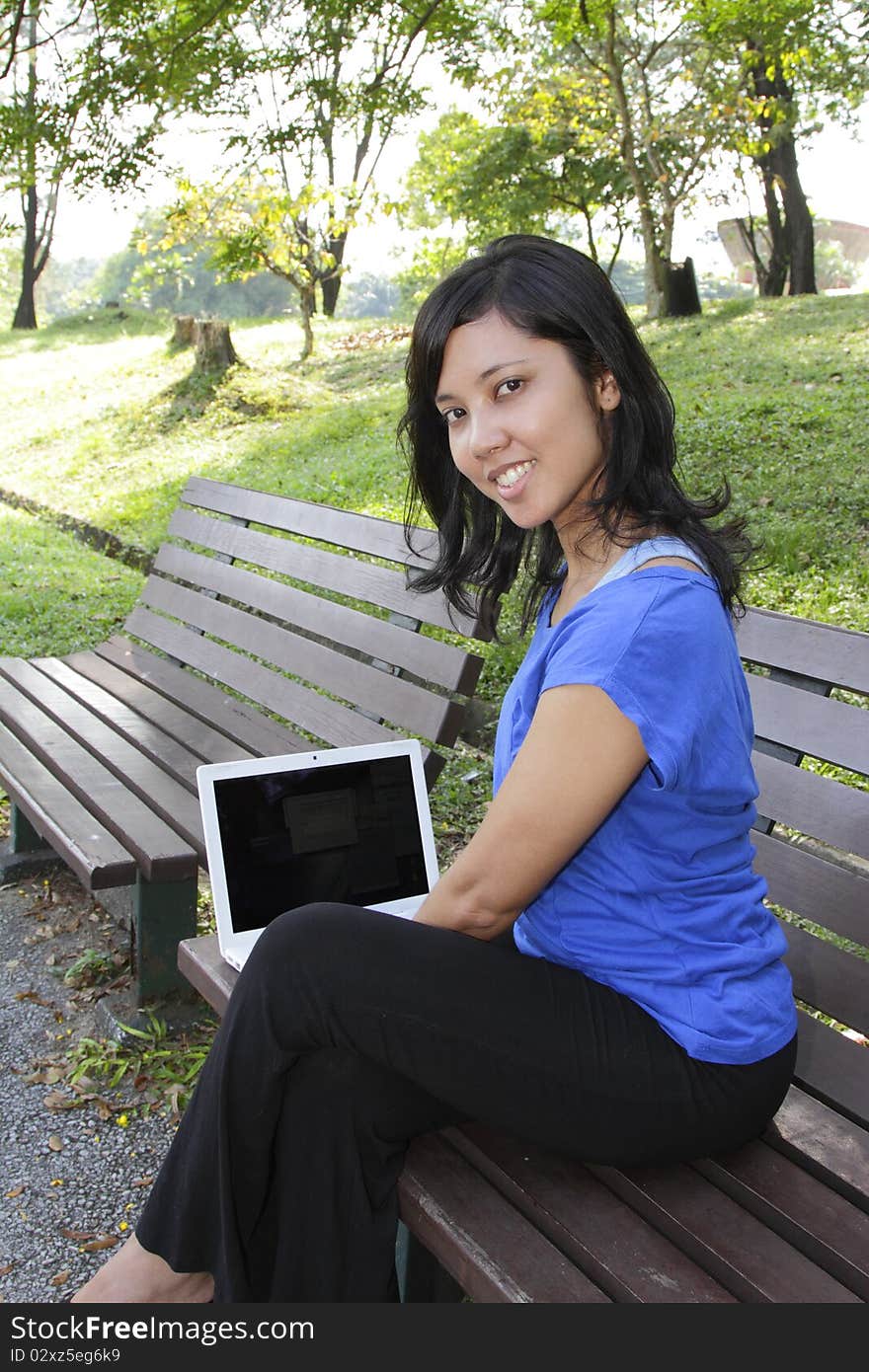 An Asian woman using a laptop on a bench at a park. An Asian woman using a laptop on a bench at a park