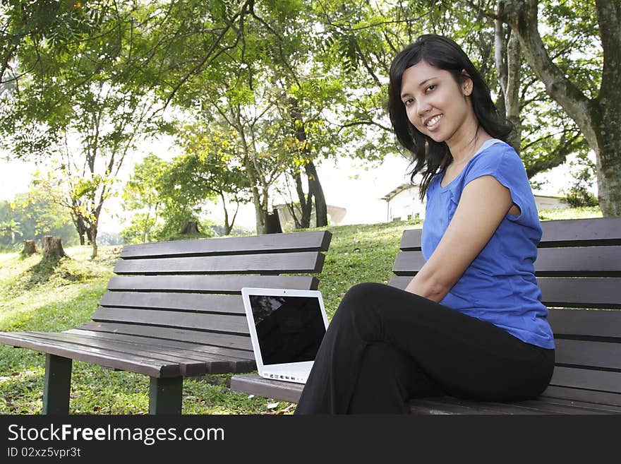 An Asian woman using a laptop on a bench at a park. An Asian woman using a laptop on a bench at a park