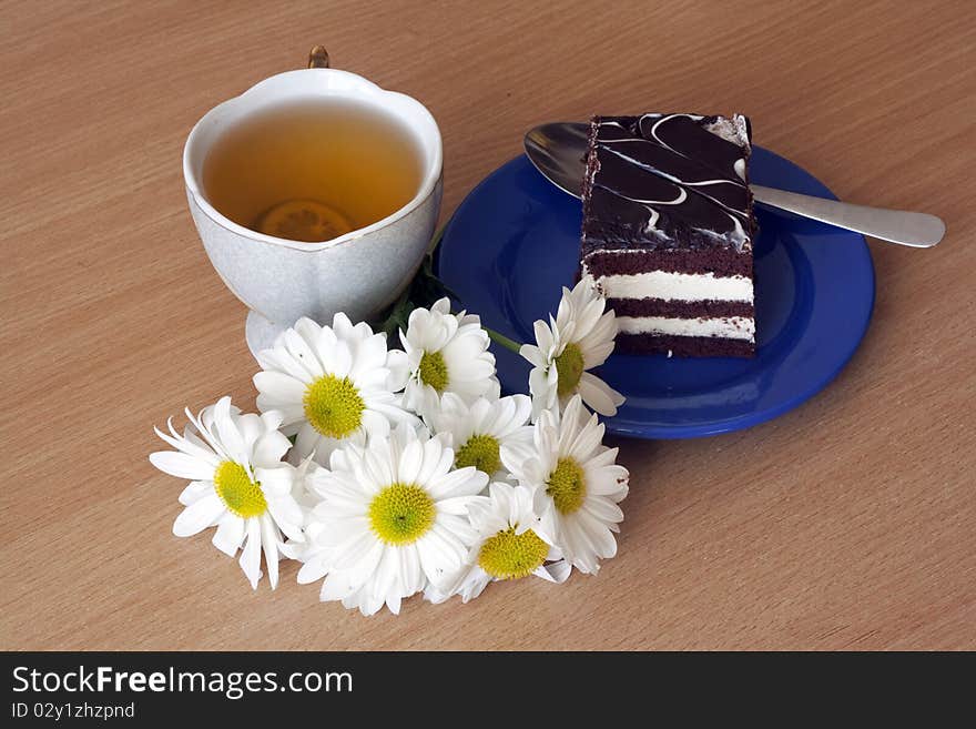 A cup of tea with lemon and a slice of chocolate cake on a plate next to a bouquet of chrysanthemums. A cup of tea with lemon and a slice of chocolate cake on a plate next to a bouquet of chrysanthemums